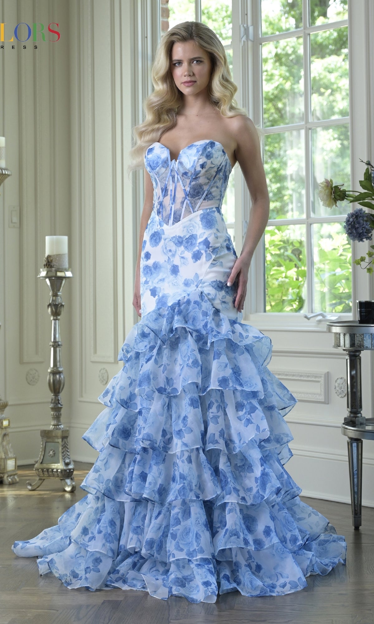 A woman stands indoors in the Long Prom Dress: Colors Dress 3928, a strapless gown with ruffled mermaid layers and a blue floral pattern, with large windows and light decor in the background.